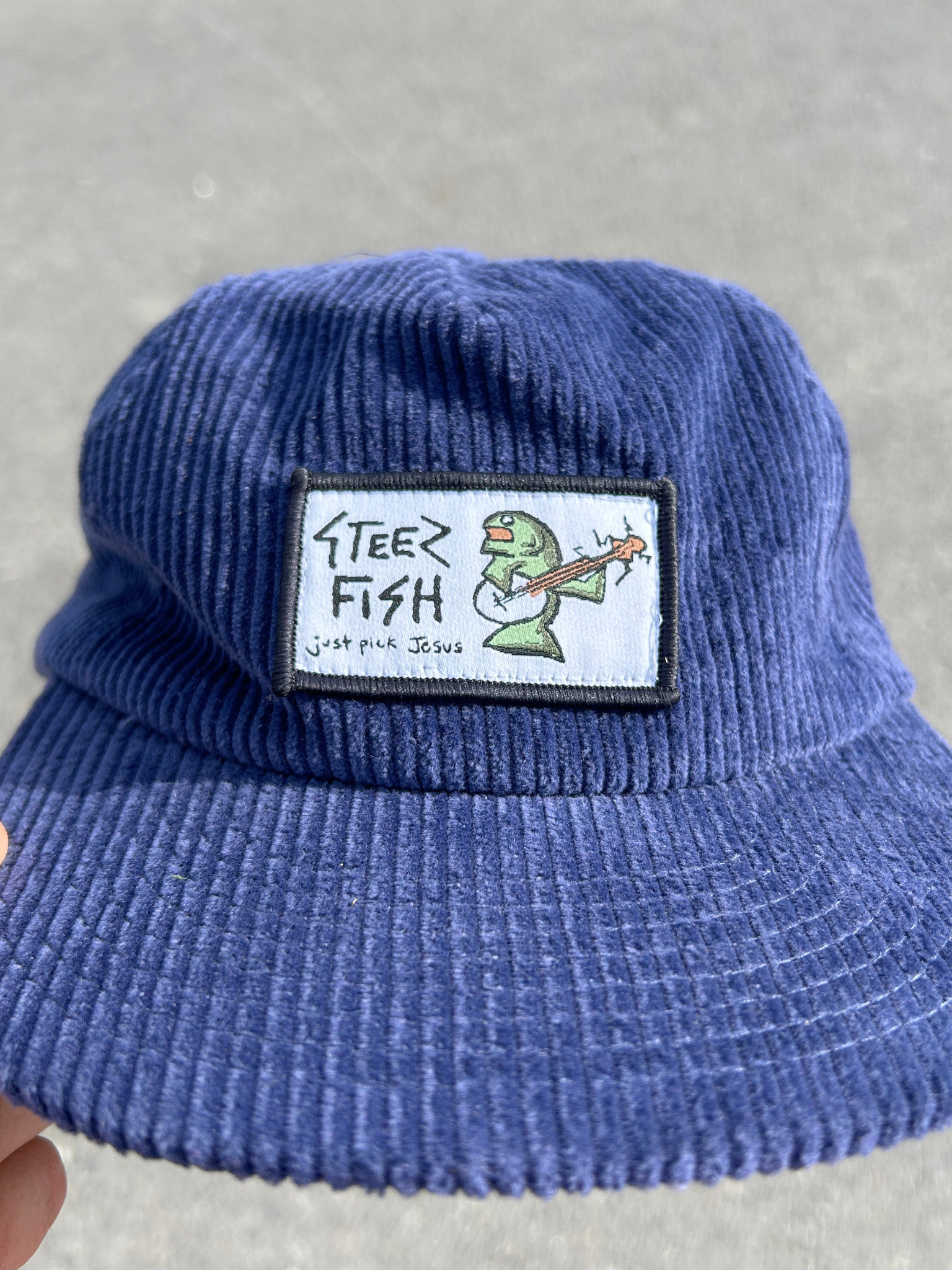 Blue corduroy hat with a patch featuring text and a fish illustration on a gray background